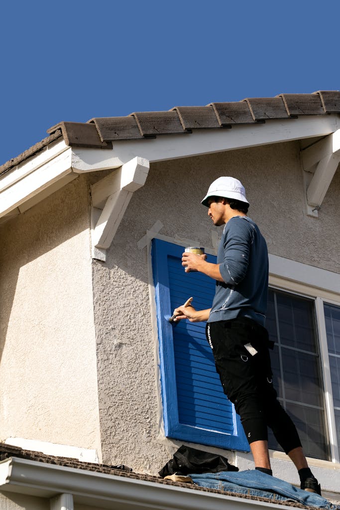 A worker painting a house shutter blue, showcasing home renovation outdoors.